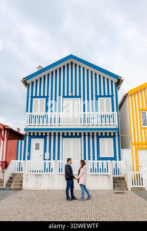 A couple stands in front of vibrant, colorful striped houses in Costa Nova, famous for their unique architectural style. The blue, red, and yellow fac Stock Photo