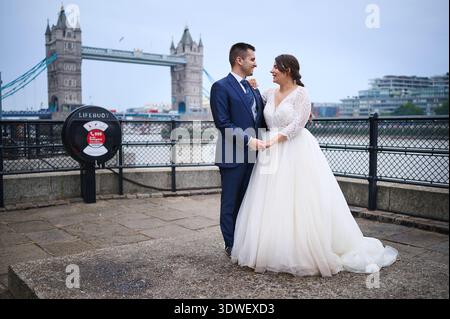 A newlywed couple poses joyfully near the iconic Tower Bridge in London. The bride in a flowing white gown and the groom in a dapper suit share a tend Stock Photo
