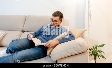 A man wearing glasses sits comfortably on a beige couch at home, reading a book. The calm atmosphere is complemented by a cozy pillow and a small indo Stock Photo