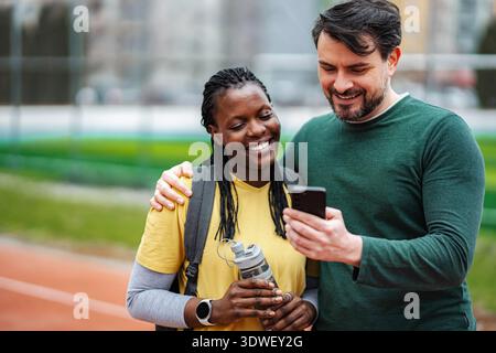 Diverse couple smiling using smartphone on running track Stock Photo