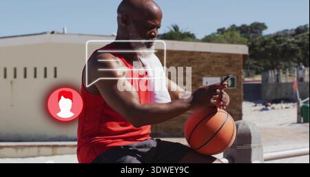 Sitting man in red tank, black shorts, towel, holding basketball, using phone on beach, chat bubble Stock Photo