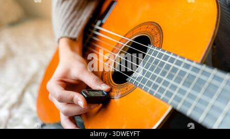 A young woman carefully tunes her classical guitar with precision, using a digital tuner to ensure perfect sound quality. The focus is on the craftsma Stock Photo