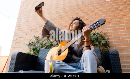 A young woman with curly hair plays an acoustic guitar while taking a selfie on a sunny patio. She wears casual attire and sits on a wicker chair agai Stock Photo
