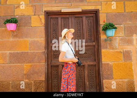 A woman with a camera explores the charming streets of Barichara, Colombia. She stands in front of a rustic wooden door, wearing a stylish outfit perf Stock Photo