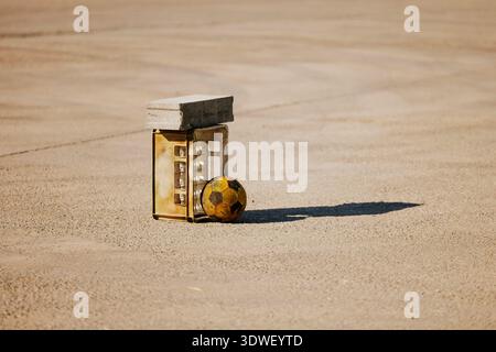 A weathered soccer ball rests against a metal crate topped with a concrete block on an expansive asphalt surface, casting a long shadow in the afterno Stock Photo