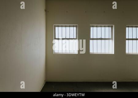 A dimly lit room with three vertical windows lined with bars. Soft natural light filters through, casting subtle shadows on the plain beige walls, evo Stock Photo