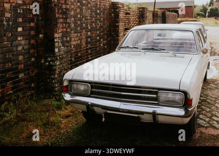 A classic white vintage car is parked alongside an aged brick wall, exuding a sense of nostalgia. The setting evokes a timeless feel, perfect for auto Stock Photo