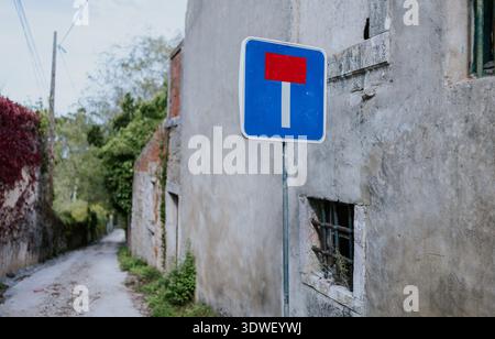 A blue and red dead-end street sign stands prominently along a narrow, rustic alley with high stone walls, lush greenery, and hints of aged architectu Stock Photo