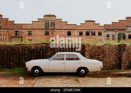A classic white car is parked in front of an abandoned brick industrial building, creating a nostalgic scene. The cobblestone street and red brick off Stock Photo