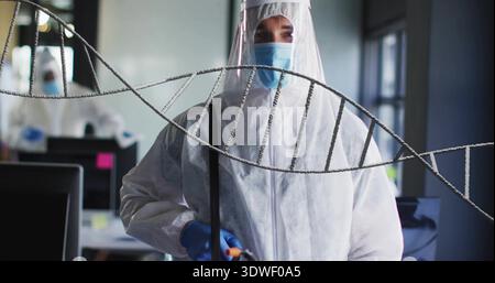 Holding probe, lab tech in coverall shield mask gloves inspecting sample in lab with helix front Stock Photo