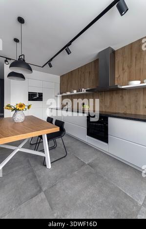 A sleek modern kitchen featuring wood-panel accents, black countertop, and white cabinets. Enhanced by track lighting and sunflowers on the table, cre Stock Photo