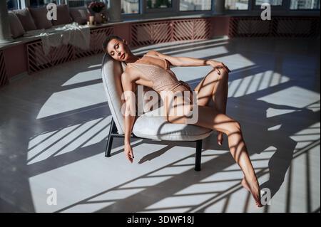 A beautiful woman relaxes in a bright studio, seated gracefully on a chair. The room is illuminated by large windows, casting deep, artistic shadows a Stock Photo
