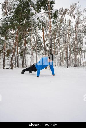 A man in a blue jacket performs push ups on a snow covered field, surrounded by tall trees. The wintry landscape underscores his dedication to outdoor Stock Photo