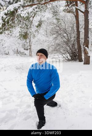 A man in a blue jacket and black beanie performs outdoor training in a snowy winter forest. The serene setting highlights dedication to fitness despit Stock Photo
