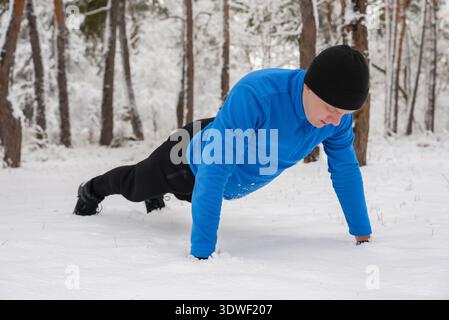A man in a blue jacket and black hat performs a push up in a snow covered forest, demonstrating dedication to outdoor fitness despite the cold winter Stock Photo