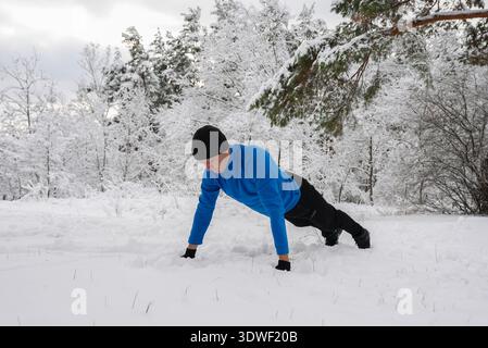 A man in a blue jacket performs push ups in a snowy winter landscape, demonstrating dedication and endurance in his outdoor fitness routine amidst a s Stock Photo