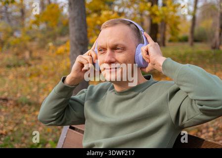 A man enjoys a peaceful moment listening to music on headphones while sitting on a park bench, surrounded by autumn foliage, reflecting leisure and re Stock Photo