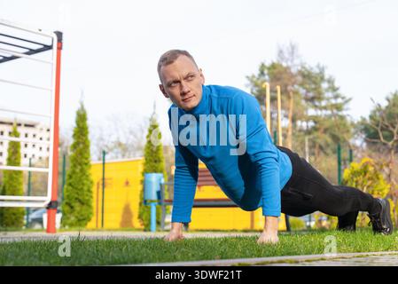 A man in a blue jacket performs outdoor exercises on grass in a park. He maintains good form with a focused expression, surrounded by green trees and Stock Photo
