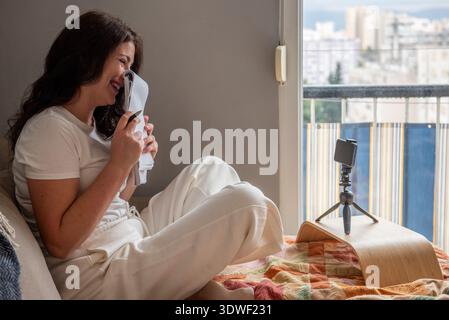 A woman is blogging from home, streaming live using her smartphone on a tripod. She is sitting comfortably, creating engaging content for her audience Stock Photo