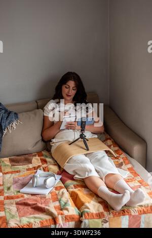 Woman sitting comfortably on a couch at home, engaged in blogging. She is live streaming using a smartphone while taking notes, with headphones and a Stock Photo