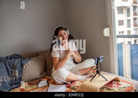 A cheerful woman sits cross-legged on a cozy couch, wearing headphones and streaming with her smartphone. She is creating engaging content for her blo Stock Photo