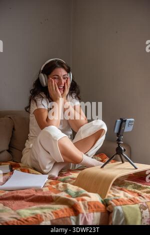 Young woman wearing headphones sits at home smiling while streaming a vlog. Using a smartphone mounted on a tripod, she engages her audience with enth Stock Photo