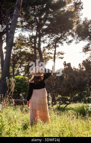 A young pregnant woman in a flowing skirt and hat stands in a sun-dappled meadow, surrounded by towering trees, capturing a serene connection with nat Stock Photo