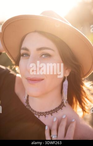 A young woman wearing a hat enjoys the warm sunlight outdoors. Her casual attire and accessories reflect a laid-back style, embraced by the sun's gold Stock Photo