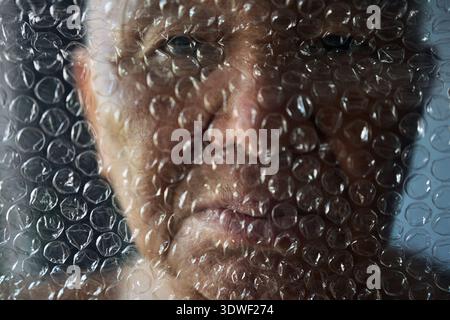 A contemplative senior man looks through clear bubble wrap. His gentle gaze and the soft texture of the material reflect themes of aging, vulnerabilit Stock Photo