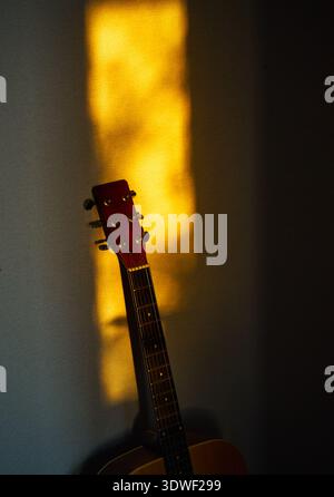 An acoustic guitar stands gracefully against a wall, bathed in the warm glow of a sunset. The soft golden light creates a peaceful and artistic atmosp Stock Photo