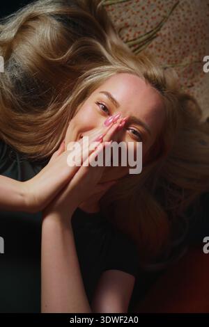 A blond woman lays back comfortably at home, her hands resting on her face. Her expression is serene and joyful, capturing a moment of relaxation and Stock Photo