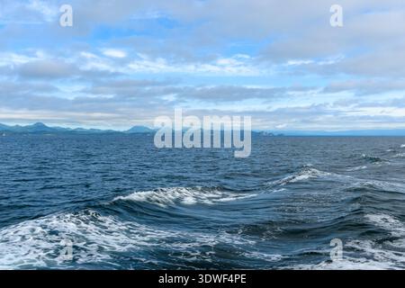 Soft waves create ripples across the ocean, while clouds drift above. Distant mountains rise against the horizon, reflecting a tranquil coastal scene. Stock Photo