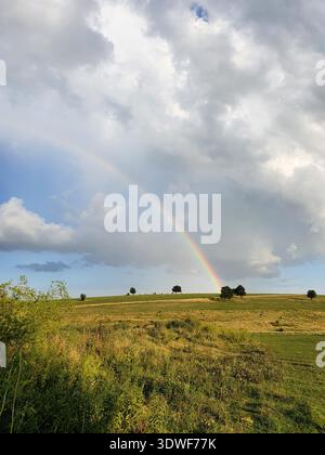 A vibrant rainbow arches across a sunlit green meadow and rolling fields beneath dramatic clouds, evoking calm, hope and natural beauty in a peaceful Stock Photo
