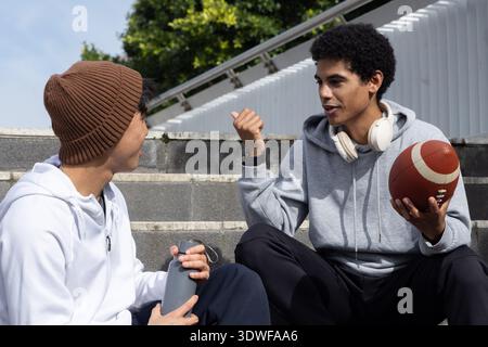 Diverse teenage male friends sitting on concrete steps at park chatting holding American football Stock Photo