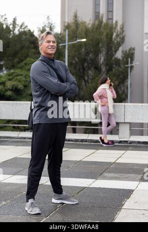 Man standing on plaza with bag and earbud, woman leaning on railing, talking on smartphone Stock Photo