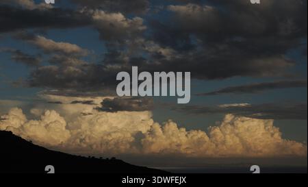Large illuminated cumulonimbus cloud formations rising above the Aegean Sea during golden hour. Dark upper clouds contrast with sunlit cloud towers, c Stock Photo