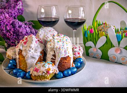 Festive Easter still life with traditional glazed cakes, two glasses of red wine, lilac blossoms, and a green grass-style basket with sweets. Stock Photo