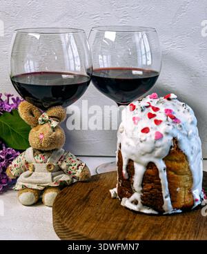 Festive Easter composition with two glasses of red wine, a cute plush bunny in a skirt, blooming lilac flowers, and colorful foil-wrapped chocolate eg Stock Photo