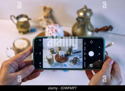 Female hands holding a smartphone to capture an Easter still life. Out-of-focus background with coffee, milk creamer, and a plush bunny in a skirt. Stock Photo