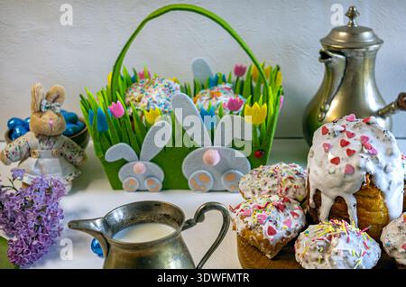 Full view of an Easter composition: a green grass-style basket with a cake, a plush bunny in a skirt, lilac flowers, a moka pot, and a milk creamer. Stock Photo
