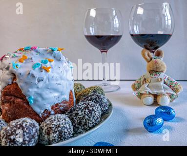 Festive Easter composition with two glasses of red wine, a cute plush bunny in a skirt, blooming lilac flowers, and colorful foil-wrapped chocolate eg Stock Photo