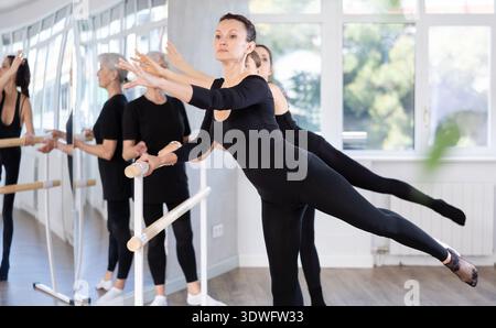 Mature teacher conducts class with girls in ballet studio, observes doing arabesque exercise. Stock Photo