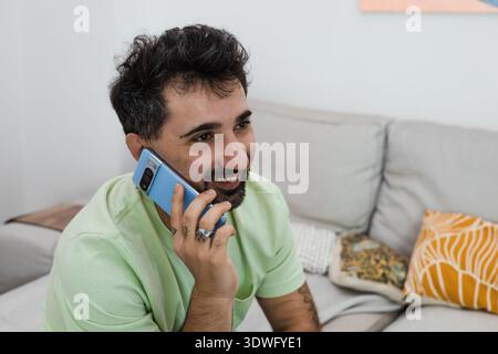 Smiling man in a mint green shirt having a relaxing conversation on his smartphone while seated on a comfortable couch with colorful cushions at home, Stock Photo