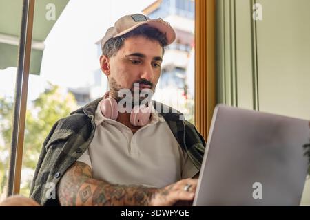 A man with a beard and tattoos, wearing headphones and a cap, focuses intently on his laptop in a brightly lit cafe environment, embodying a modern, c Stock Photo