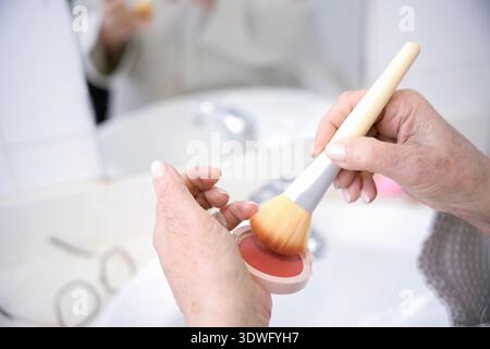 A close-up image of senior hands applying blush with a soft makeup brush. The scene captures the delicate touch of the brush on the blush in a bathroo Stock Photo