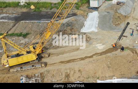 A large yellow crane at an active construction site moving materials while two workers in orange safety vests collaborate on the ground. Dirt and mach Stock Photo