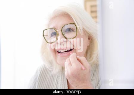 An elderly woman smiling joyfully while applying lipstick, wearing large glasses, and dressed in a cozy robe, embodying a sense of happiness, confiden Stock Photo