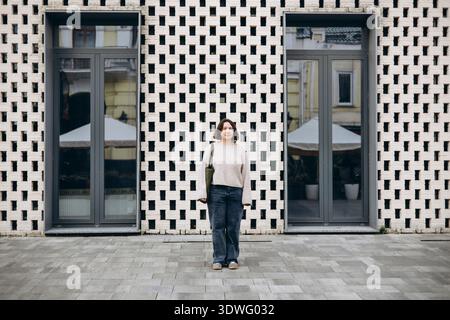 A woman stands in front of a patterned brick wall with two large glass windows reflecting the urban landscape. She carries a shoulder bag and wears ca Stock Photo