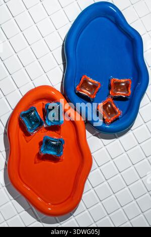 Vibrant photo of laundry capsules placed on colorful orange and blue dishes against a white tiled background, showcasing the balance of aesthetic and Stock Photo