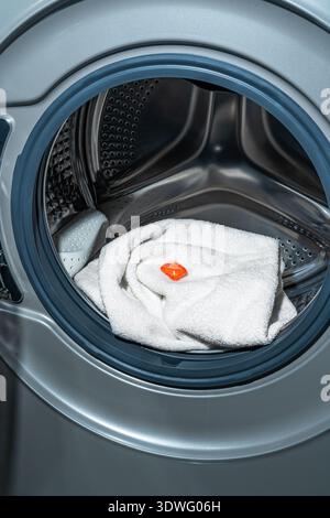 A vibrant orange laundry capsule sits atop a neatly folded white towel inside a modern front-loading washing machine, ready for a washing cycle. Perfe Stock Photo